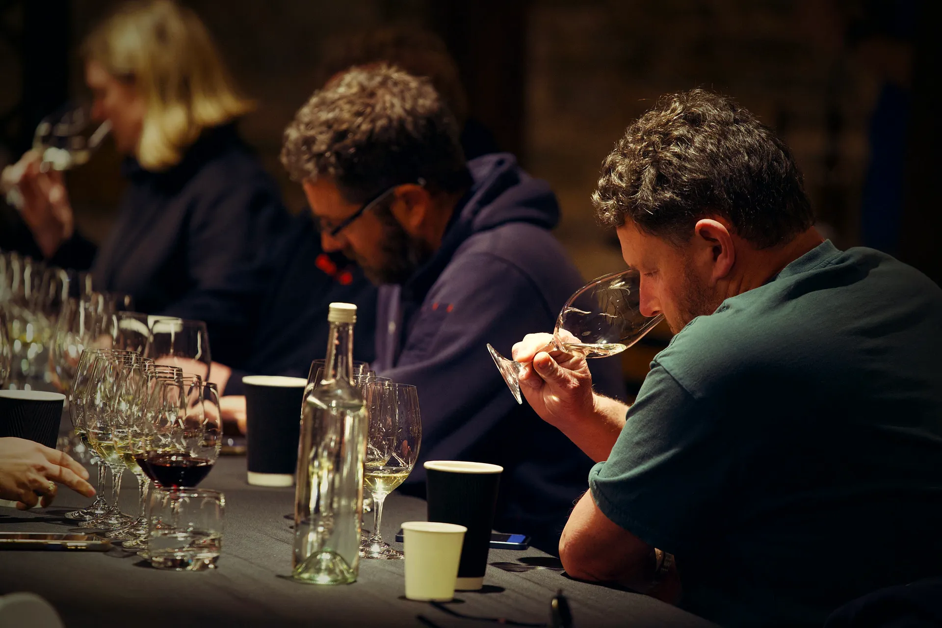 A row of competitors at the long table, each nose-deep in a tilted glass, working a question.
