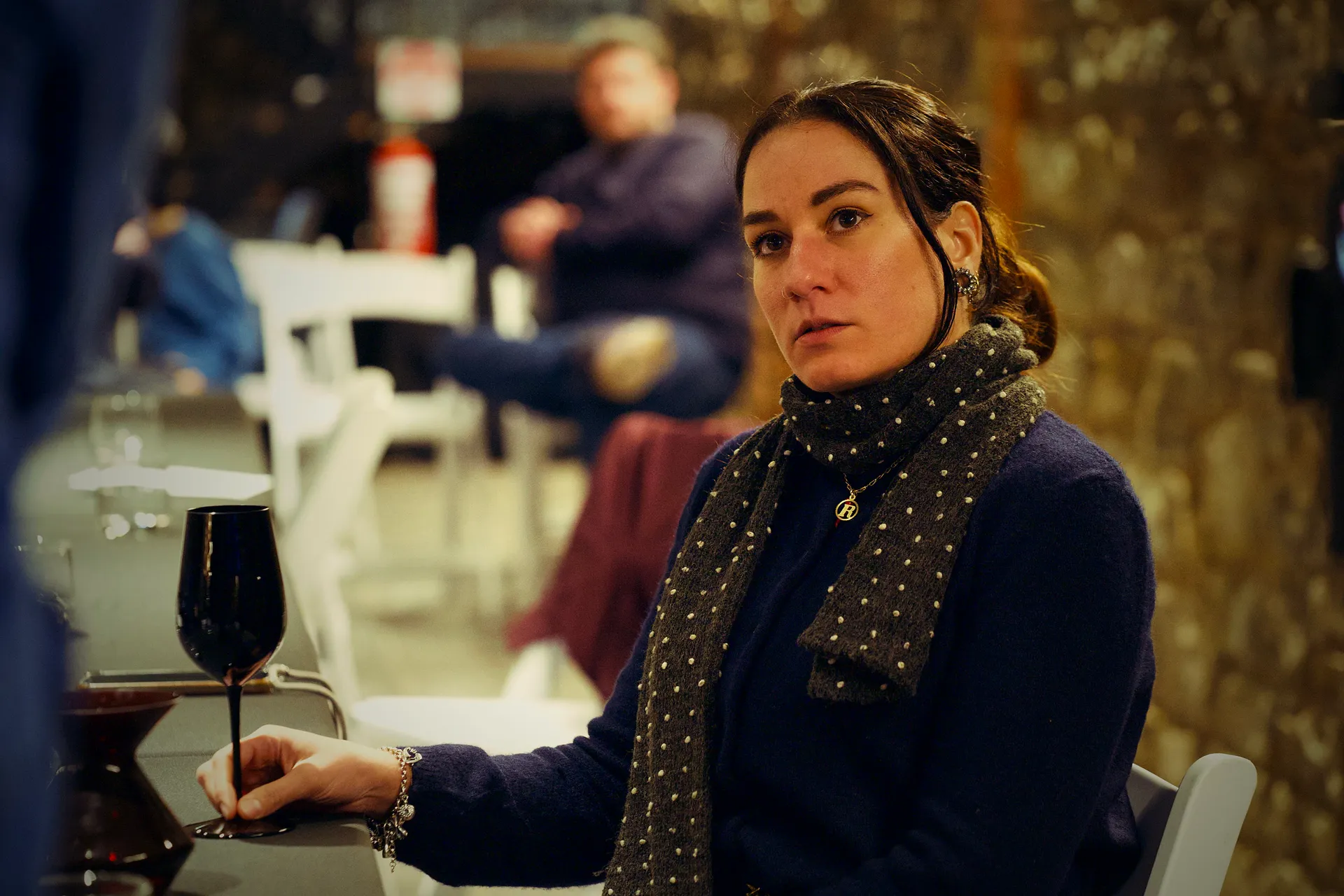 Roberta Ruotolo at the table, hand on the stem of a black glass, decanter beside her, eyes on the front of the room.