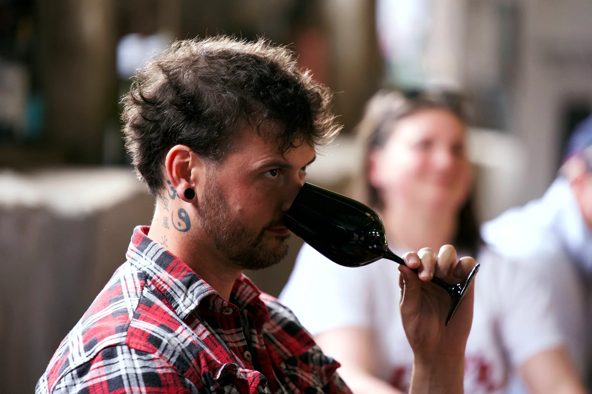 A player in a red checked shirt noses a black glass, eyes to one side, while someone smiles in the soft background.